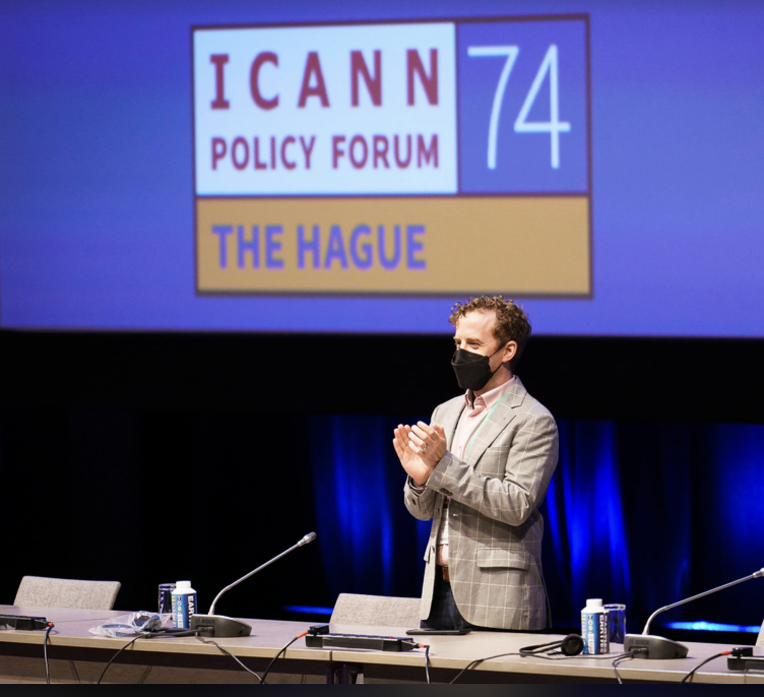 Person in a suit and mask standing and clapping at a conference with an ICANN Policy Forum sign in the background.